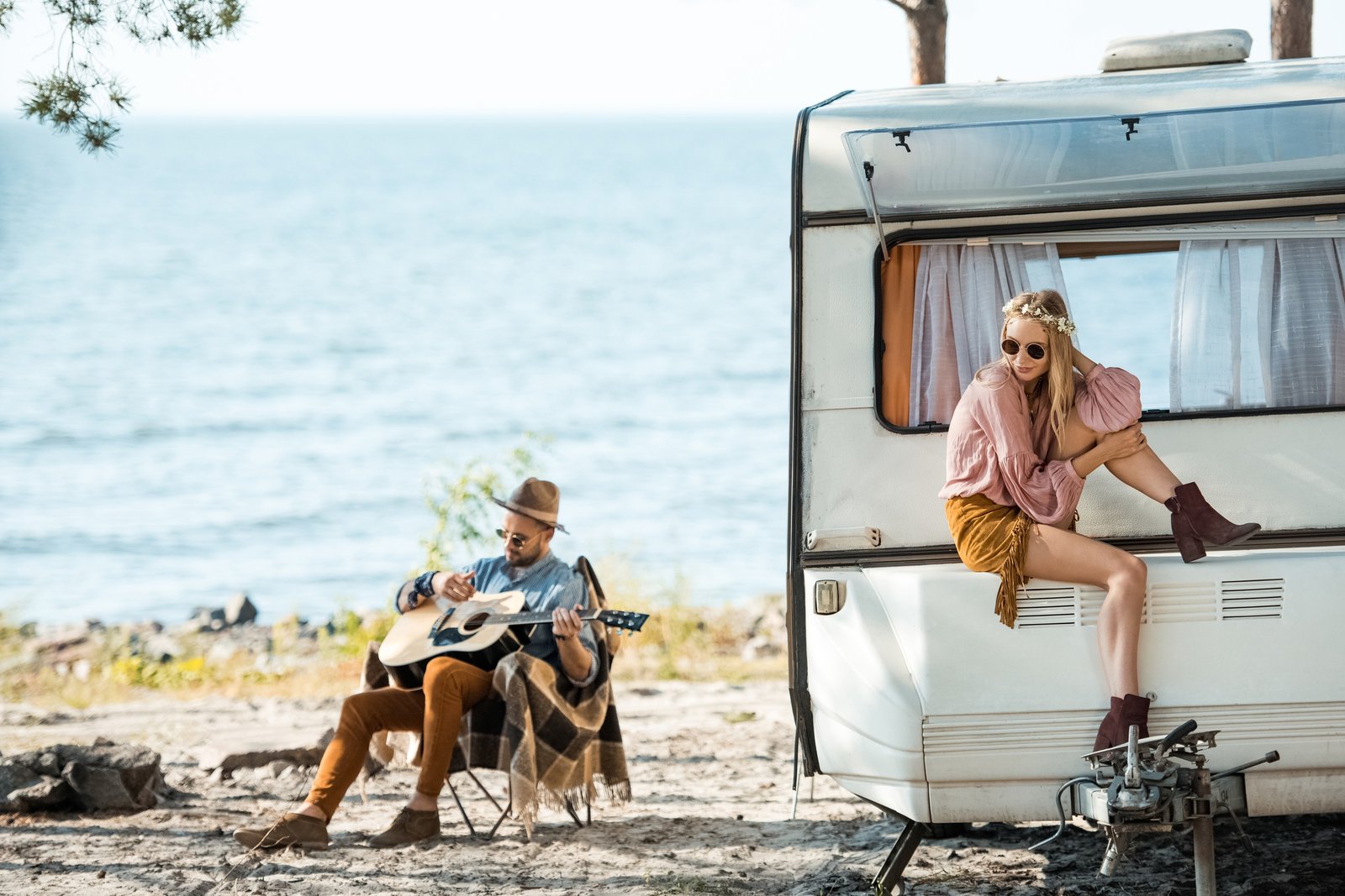 beautiful hippie girl sitting on campervan while man playing guitar near sea