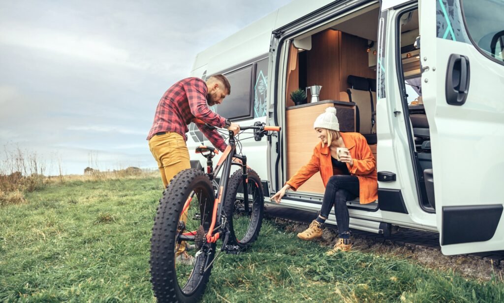 Woman drinking coffee sitting at the door of a campervan and man checking fat bike