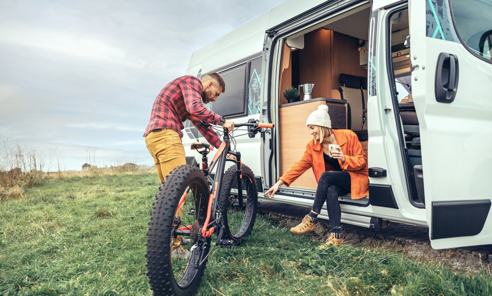 Woman drinking coffee sitting at the door of a campervan and man checking fat bike